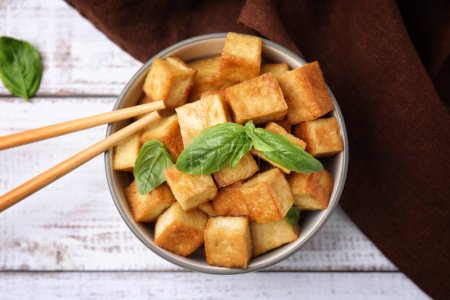 Bowl with delicious fried tofu, basil on white wooden table, flat lay