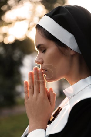 Téléchargez les photos : Jeune religieuse avec les mains serrées ensemble priant à l'extérieur le jour ensoleillé - en image libre de droit