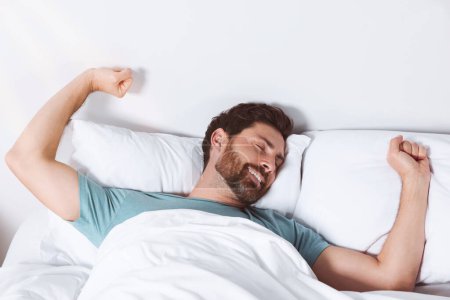 Téléchargez les photos : Homme heureux s'étirant sur des oreillers confortables dans le lit à la maison - en image libre de droit