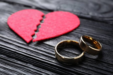 Divorce concept. Broken red paper heart and wedding rings on black wooden table, closeup