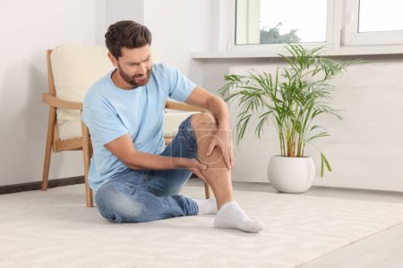 Man suffering from leg pain on white carpet near armchair at home