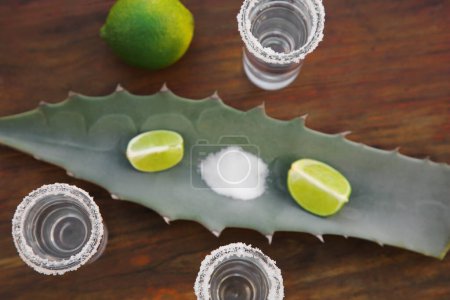 Foto de Tiros de tequila mexicano, sal, lima y hoja verde sobre mesa de madera, puesta plana. Bebida hecha de agava - Imagen libre de derechos