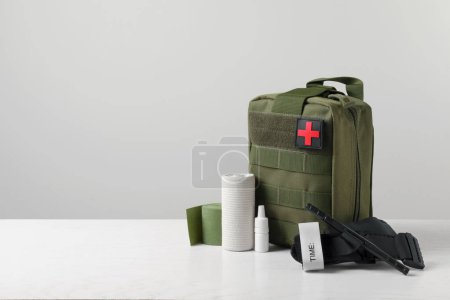Botiquín militar de primeros auxilios, torniquete, gotas y venda elástica sobre mesa blanca, espacio para texto