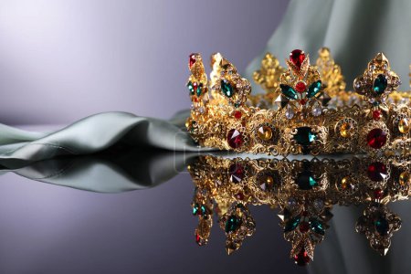 Téléchargez les photos : Beautiful golden crown with gems near light cloth on dark mirror surface. Space for text - en image libre de droit