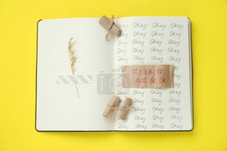 Photo for Phrase It`s Ok to Not Be Ok, rolled pieces of paper and dry flower attached with adhesive tape in notebook on yellow background, top view - Royalty Free Image