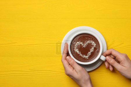 Téléchargez les photos : Femme tenant tasse de chocolat chaud avec décoration en forme de coeur à la table en bois jaune, vue de dessus. Espace pour le texte - en image libre de droit
