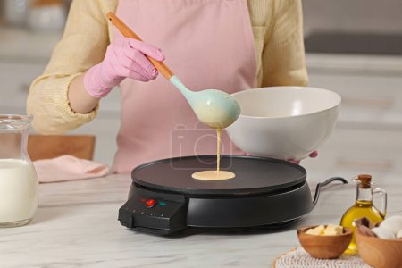 Woman cooking delicious crepe on electric pancake maker at white marble table in kitchen, closeup