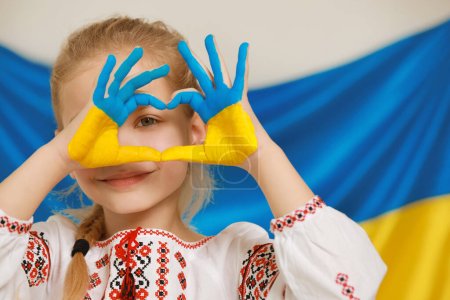 Kleines Mädchen bastelt Herz mit bemalten Händen in der Nähe der ukrainischen Flagge, Raum für Text. Liebe Ukraine-Konzept