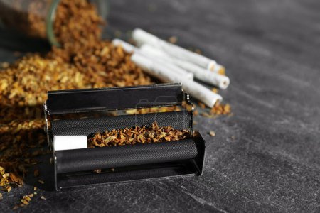 Roller, tobacco and hand rolled cigarettes on dark grey table, closeup. Space for text