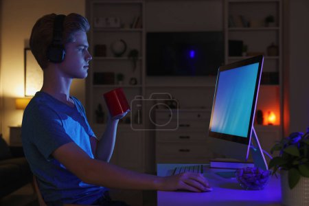 Teenage boy with cup of drink using computer in room at night. Internet addiction Poster 645729028