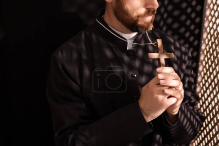 Foto de Sacerdote católico en sotana sosteniendo cruz en cabina confesional, primer plano - Imagen libre de derechos
