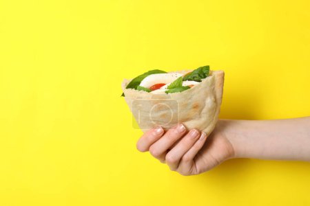 Woman holding delicious pita sandwich with mozzarella, tomatoes and basil on yellow background, closeup