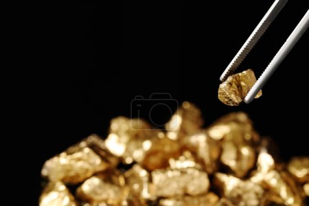 Tweezers with gold nugget against blurred background, closeup. Space for text