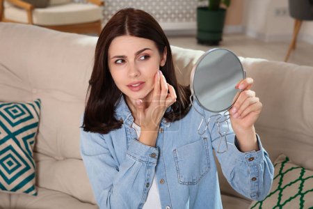 Young woman looking in mirror and squeezing pimple indoors. Hormonal disorders
