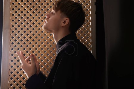 Téléchargez les photos : Prêtre catholique priant près d'une fenêtre en bois dans une cabine confessionnelle - en image libre de droit