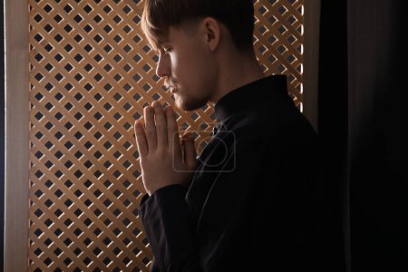 Foto de Sacerdote católico rezando cerca de ventana de madera en cabina confesional - Imagen libre de derechos