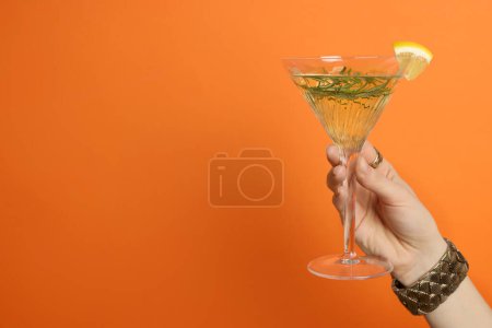 Woman holding martini glass of refreshing cocktail with lemon slice and rosemary on orange background, closeup. Space for text