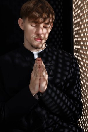 Foto de Sacerdote católico rezando cerca de ventana de madera en cabina confesional - Imagen libre de derechos