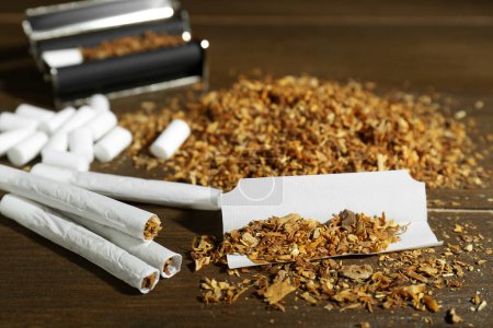 Tobacco, hand rolled cigarettes and roller on wooden table, closeup