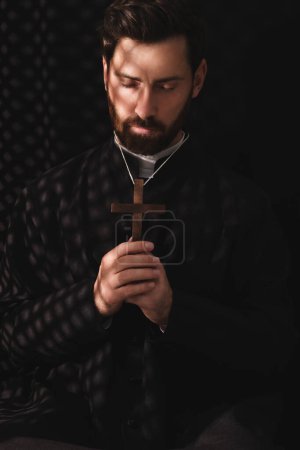 Foto de Sacerdote católico en sotana sosteniendo la cruz y orando a Dios en confesionario - Imagen libre de derechos