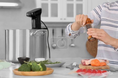 Woman grinding pepper onto meat near pot with sous vide cooker in kitchen, closeup. Thermal immersion circulator