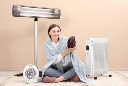 Jeune femme avec différents appareils de chauffage électriques modernes près du mur beige à l'intérieur