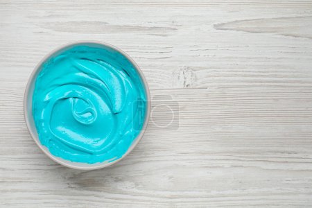 Téléchargez les photos : Bol de crème avec colorant alimentaire bleu clair sur table en bois blanc, vue de dessus. Espace pour le texte - en image libre de droit