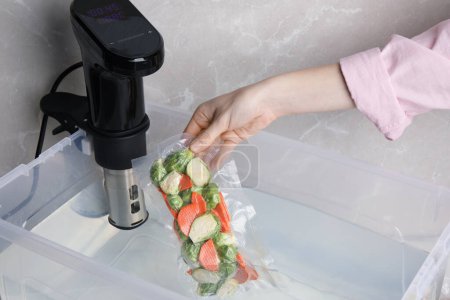 Foto de Woman putting vacuum packed vegetables into box with thermal immersion circulator, closeup. Sous vide cooking - Imagen libre de derechos