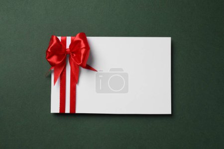 Blank gift card with red bow on dark green background, top view. Space for text