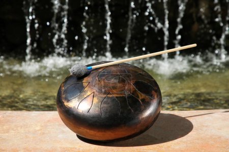 Steel tongue drum with mallet near waterfall outdoors on sunny day. Percussion musical instrument