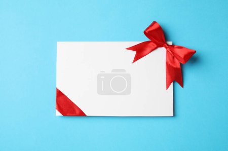 Blank gift card with red bow on light blue background, top view. Space for text