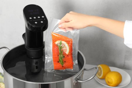 Photo for Woman putting vacuum packed salmon into pot with sous vide cooker in kitchen, closeup. Thermal immersion circulator - Royalty Free Image