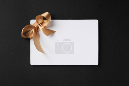 Téléchargez les photos : Carte cadeau vierge avec noeud doré sur fond noir, vue de dessus. Espace pour le texte - en image libre de droit