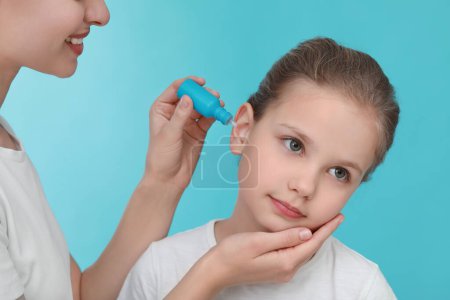 Téléchargez les photos : Mère goutte à goutte des médicaments dans l'oreille de sa fille sur fond bleu clair - en image libre de droit