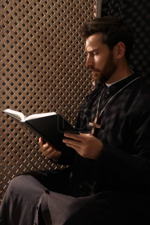 Foto de Sacerdote católico en sotana leyendo la Biblia en cabina confesional - Imagen libre de derechos