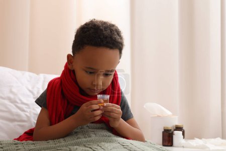 Foto de Niño afroamericano tomando jarabe para la tos en la cama en casa, espacio para el texto. Medicina para el resfriado - Imagen libre de derechos