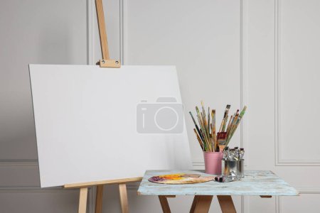 Foto de Caballete con lienzo en blanco y diferentes suministros de arte en la mesa de madera cerca de la pared blanca - Imagen libre de derechos