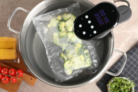 Thermal immersion circulator and vacuum packed broccoli in pot on grey marble table, flat lay. Sous vide cooking