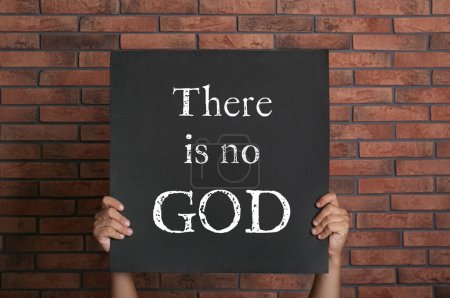 Photo for Atheism. Man holding sign with text There Is No God near brick wall, closeup - Royalty Free Image