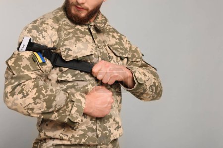Foto de Soldado ucraniano en uniforme militar aplicando torniquete médico en el brazo sobre fondo gris claro, primer plano. Espacio para texto - Imagen libre de derechos