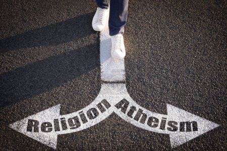 Foto de Elección entre ateísmo y religión. Hombre caminando hacia marcas dibujadas en la carretera, primer plano. Flechas con palabras apuntando en direcciones opuestas - Imagen libre de derechos
