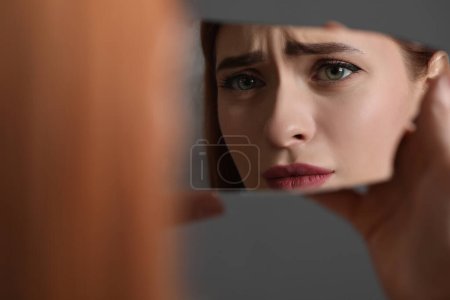 Photo for Young woman looking at herself in shard of broken mirror on grey background, closeup - Royalty Free Image