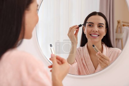 Beautiful young woman applying eyebrow gel near mirror indoors
