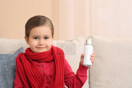 Cute little girl holding nasal spray on sofa indoors