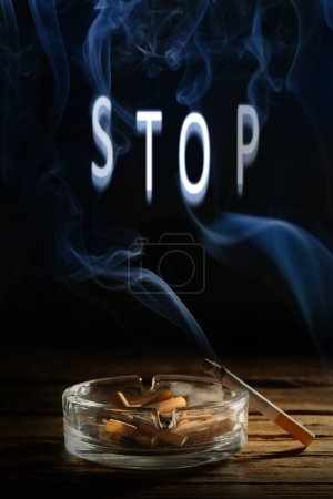 Photo for Quit smoking. Word Stop of smoke, ashtray with stubs and smoldering cigarette on wooden table against black background - Royalty Free Image