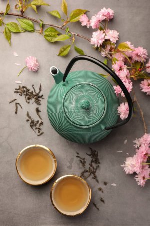 Traditional ceremony. Cups of brewed tea, teapot and sakura flowers on grey table, flat lay
