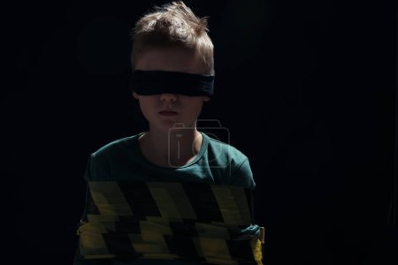 Photo for Blindfolded little boy tied up and taken hostage against dark background - Royalty Free Image