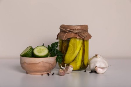 Foto de Sabrosos pepinos en escabeche, verduras cortadas en un tazón de madera y ajo en una mesa blanca - Imagen libre de derechos