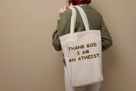 Woman holding bag with phrase Thank God I Am Atheist on beige background, closeup