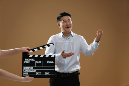 Happy asian actor performing while second assistant camera holding clapperboard on brown background. Film industry